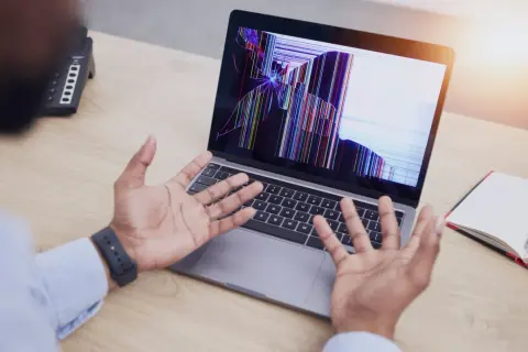 Rozlany ekran w laptopie wyświetla kolorowe pęknięcia, a osoba z rozłożonymi rękami patrzy na niego z niedowierzaniem.
