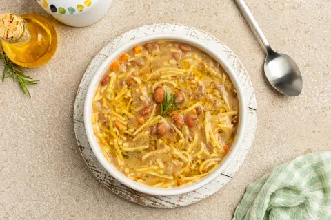 Un bol de pasta e fagioli, une soupe réconfortante avec des pâtes fines et des haricots, garnie de romarin. Une cuillère et une petite bouteille d'huile d'olive sont à proximité.