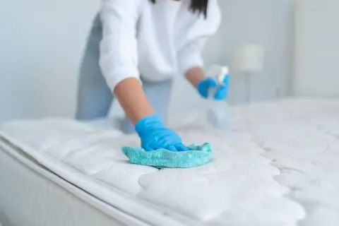 Person mit blauen Handschuhen reinigt die Matratze mit einem Tuch und Sprühflasche.