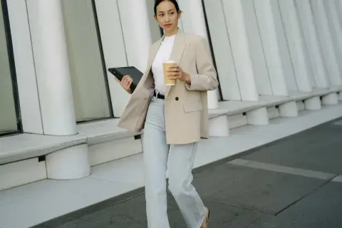 Eine Frau im smart casual Look mit beigem Blazer, Jeans und Kaffee in der Hand, geht mit einem Tablet.