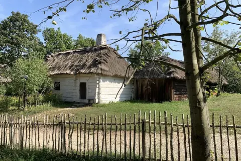Drewniane chaty kryte strzechą w skansenie, przed nimi płotek z gałęzi.