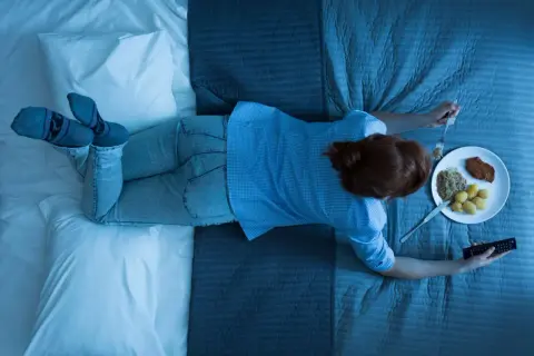 A woman eats dinner in bed, holding a remote. Wondering how long should you wait to sleep after eating?