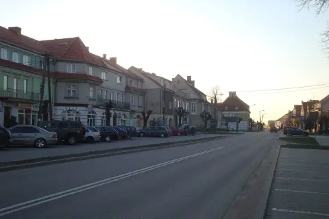 Ulica w miasteczku, gdzie są Wydminy. Rząd kamienic z czerwonymi dachami, zaparkowane samochody i droga.