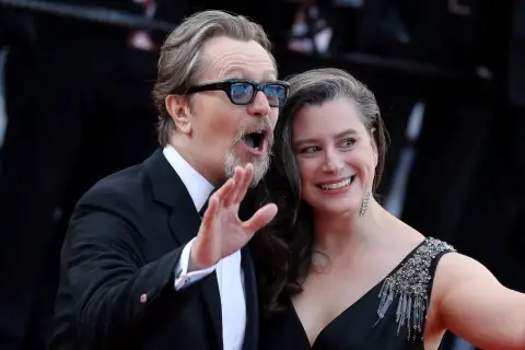 Gary Oldman z żoną, uśmiechnięci i pełni życia, pozują do zdjęć na czerwonym dywanie.