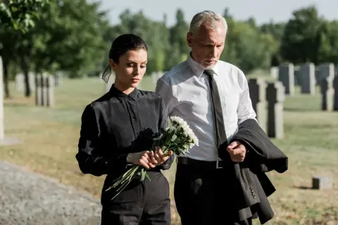 Para idąca przez cmentarz, kobieta w czerni z bukietem kwiatów, mężczyzna w białej koszuli z marynarką.