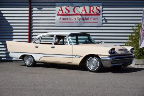 En krämvit och ljusbeige De Soto från 1950-talet står parkerad framför en byggnad med skylten "AS CARS".
