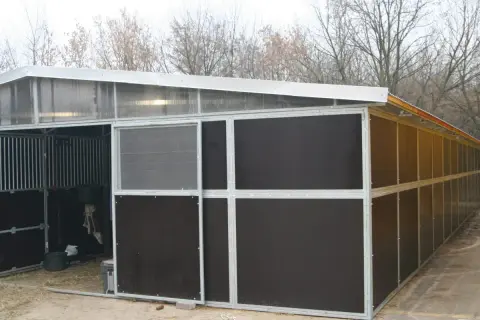 Nowa stajnia dla kucyka, z metalową konstrukcją i ciemnymi panelami. Widać kraty boksów i przezroczysty dach.