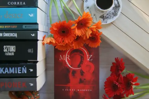 Stos książek, pomarańczowe gerbery i kubek kawy. Na okładce jednej z nich widnieje "Czarna Madonna" i tytuł "Mróz".