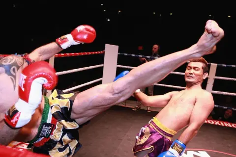 A fighter's leg swings high in a Muay Thai match. The opponent's flinching reflex is evident as he attempts to block the powerful kick.