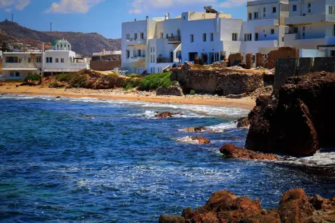 Blågrönt hav sköljer mot klippor och sandstrand. Vita hus med gröna tak reser sig mot en bergig bakgrund på Naxos, Grekland.