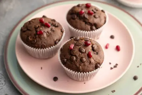 Drei saftige, vegane Muffins mit Schokostückchen und Granatapfelkernen auf einem rosa Teller.