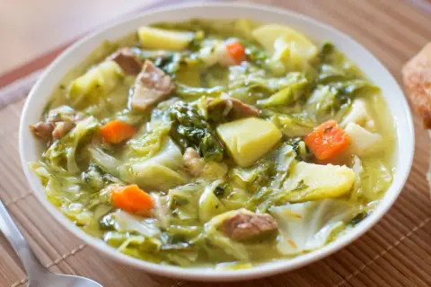 Bol de soupe au chou vert, pommes de terre, carottes et viande, avec du pain à côté.