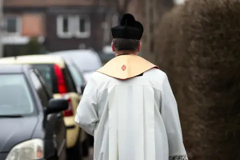 Ksiądz w sutannie i birecie odchodzi od samochodów. Co się mówi do księdza na dowidzenia? "Szczęść Boże!
