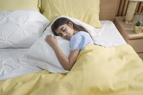 A young girl sleeps peacefully under a yellow blanket. This serene scene reminds us of the importance of fresh air and how to get rid of off-gassing smells for a healthy sleep environment.