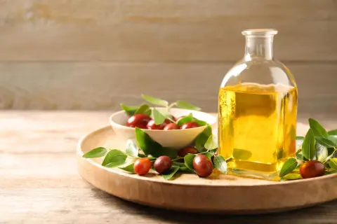 Flasche mit goldenem Simmondsia Chinensis Seed Oil neben einer Schale mit Jojobasamen und grünen Blättern auf einem Holzteller.