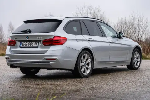 Srebrne BMW Seria 3 kombi z polskimi tablicami rejestracyjnymi stoi na mokrej nawierzchni, otoczone suchą trawą i nagimi drzewami.