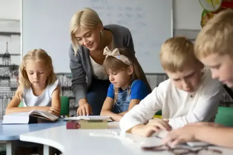 Nauczycielka wspiera uczniów w nauce, pokazując swoje kompetencje rady pedagogicznej. Dzieci skupione na książkach.
