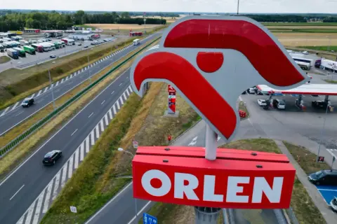 Duży znak ORLEN nad stacją paliw przy autostradzie. W tle parking z ciężarówkami i samochodami.