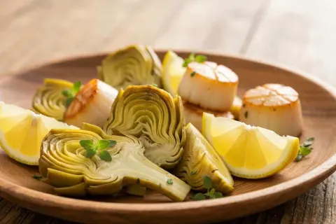 Assiette de Saint-Jacques poêlées et fleur d'artichaut coupée, garnie de quartiers de citron et de quelques herbes fraîches.