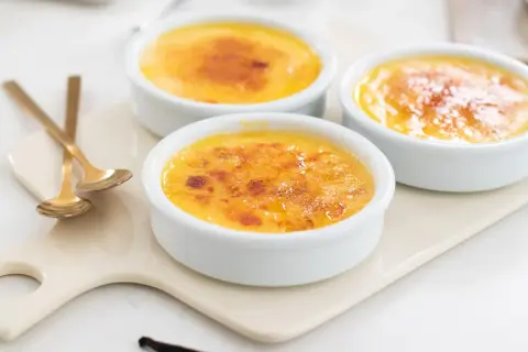 Tre krämiga creme brulee i vita skålar med karamelliserat sockerlock. Guldfärgade skedar ligger bredvid.
