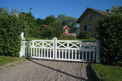 Vit grind med korsmönster leder in på en grusgång entre till ett gult hus och en röd stuga.