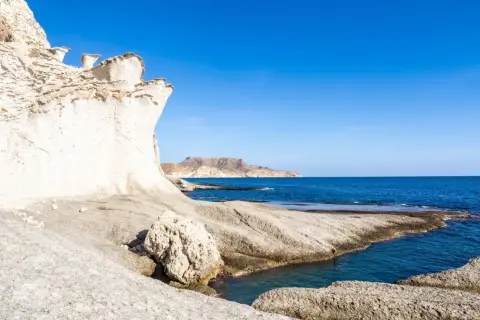 Formaciones rocosas blancas y erosionadas en una cala blanca Almería, con el mar azul intenso y montañas al fondo.