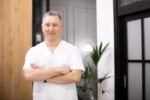 Uśmiechnięty lekarz w białej koszuli z rękami skrzyżowanymi na piersi, stoi w nowoczesnym wnętrzu.
