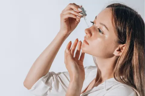 Frauenhand trägt Tropfen eines Serums auf das Gesicht auf. Ob Squalan oder Hyaluron, die Haut strahlt Feuchtigkeit aus.