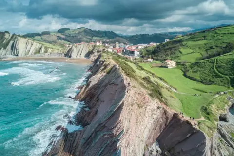 Vacker kustlinje med dramatiska klippor och gröna kullar i **Galicien, Spanien**. Havet sköljer mot stranden.