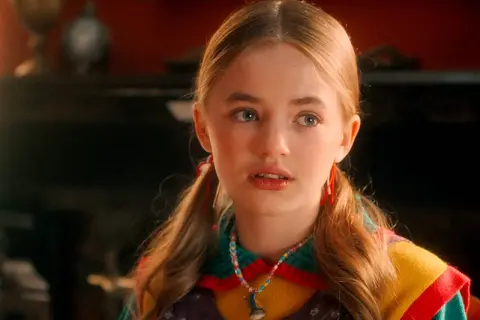 Holly Wheeler aus Stranger Things mit Zöpfen und einer bunten Halskette.