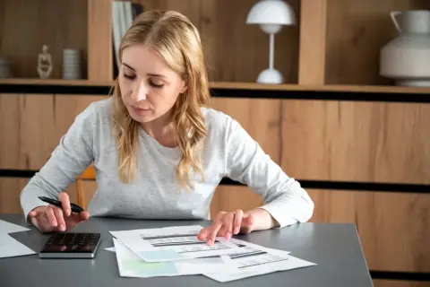 Kobieta z kalkulatorem i dokumentami analizuje wydatki, być może w celu uzyskania dodatku mieszkaniowego.