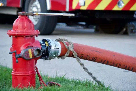 Czerwony hydrant naziemny podłączony do węża strażackiego, z tyłu widać fragment wozu strażackiego.
