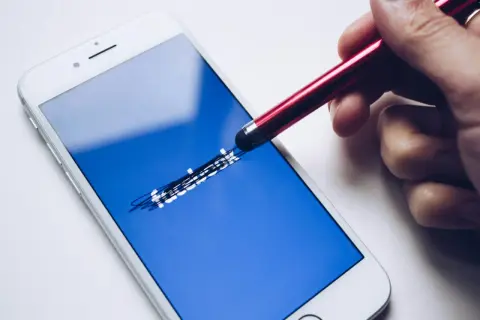 Dłoń z rysikiem próbuje sprawdzić, jak sprawdzić ile zostało blokady na FB, dotykając ekranu telefonu z logo Facebooka.