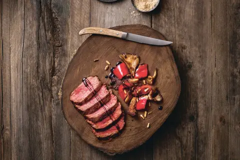 Saftige Tagliata di Manzo mit gebratenen Pilzen und Paprika auf einem rustikalen Holzbrett, garniert mit Pinienkernen und Balsamico.