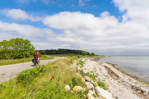 En cyklist njuter av en vacker dag längs en kustväg, en av många cykelleder i Danmark. Havet glittrar bredvid den steniga stranden.
