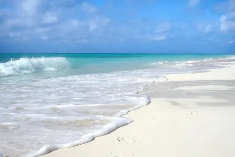 Onda che lambisce la sabbia bianca di una spiaggia d'inverno, con cielo azzurro e nuvole sparse.