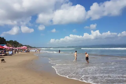 Słoneczny dzień na plaży w Seminyak na Bali. Ludzie spacerują po piasku i kąpią się w oceanie.