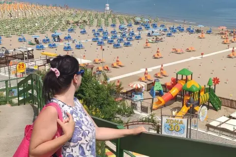 Donna con borsa rosa ammira la spiaggia di Gabicce Mare centro, con ombrelloni, lettini e un parco giochi colorato.