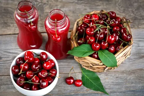 Dwa szklane dzbanki z rubinowym sokiem z czereśni, obok koszyk pełen świeżych owoców i miseczka z czereśniami.