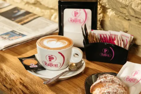Un cappuccino senza schiuma, con un cuore di latte art, accompagnato da un croissant e un giornale.