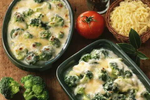 Deux plats de gratin de brocolis crémeux et gratinés, accompagnés de brocolis frais, d'une tomate et de fromage râpé.