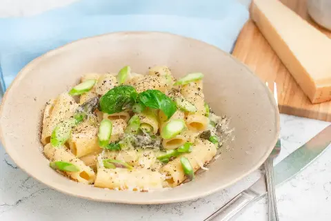 En krämig tallrik med pasta med sparris, toppad med färsk basilika och riven parmesan. En bit parmesan ligger bredvid.