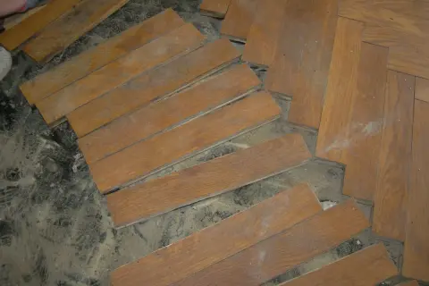 Drewniana podłoga w jodełkę, częściowo rozebrana. Widać pojedyncze deseczki i fragmenty ułożonego parkietu. Subit co to?