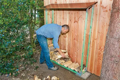 Mann lagert brennholz unter einem Holzunterstand. Der Untergrund ist trocken und schützt das Holz vor Feuchtigkeit.