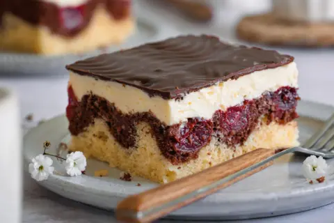 Ein Stück Donauwelle-Kuchen mit Schokoladenglasur, Sahne und Kirschen. Ein köstliches Rezept für den perfekten Genuss.
