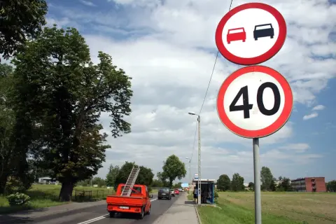 Znak zakazu wyprzedzania i ograniczenie prędkości do 40 km/h. Pomarańczowy pick-up z drabiną jedzie po drodze.