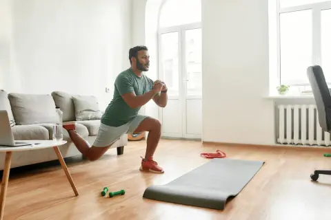 Un homme fait des fentes dans son salon, une idée pour comment commencer le sport à la maison. Un tapis, des haltères et un ordinateur sont à proximité.