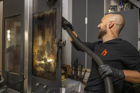 Un homme nettoie la vitre d'un poêle avec un aspirateur. Un bon entretien garantit la bonne fréquence de ramonage.