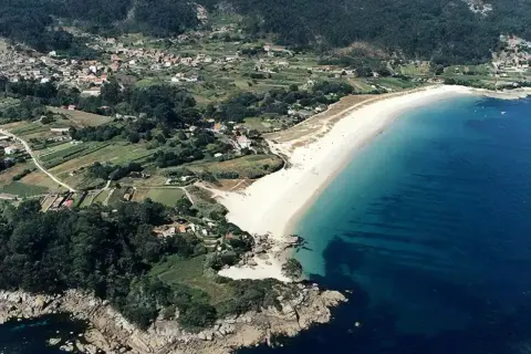 Vista aérea de la playa Area Brava, con arena blanca, aguas turquesas y un pueblo costero rodeado de vegetación y campos verdes.
