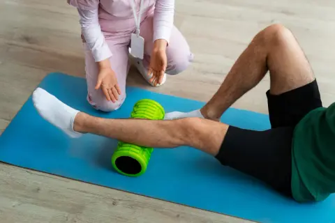Fizjoterapeuta pomaga pacjentowi w ćwiczeniach po endoprotezie kolana, używając wałka do masażu.